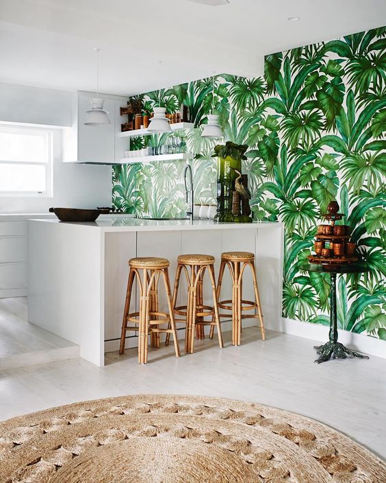 A white tropical kitchen with a tropical leaf patterned wall, rattan stools, and a jute rug looks modern and boho