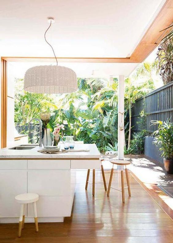 simple white kitchen island, a white wicker lampshade, wooden stools and a tropical garden that the kitchen opens onto