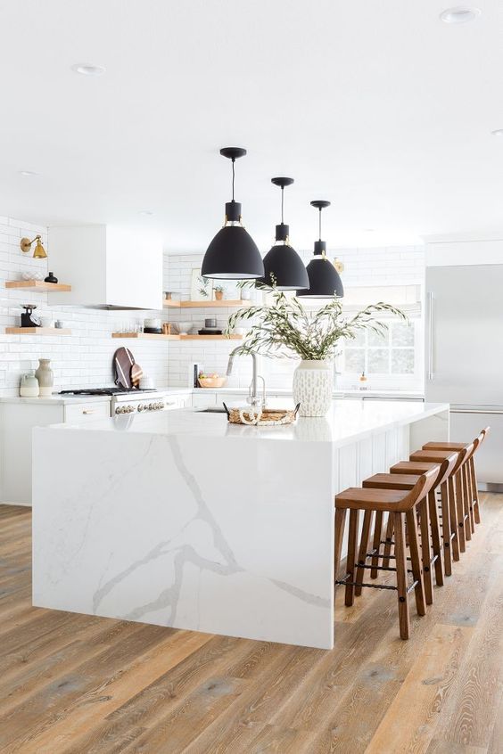 a beautiful all-white kitchen with a large island and waterfall countertop, chic black pendant lamps and wooden stools