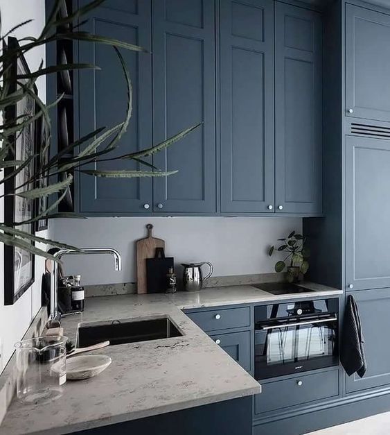 a chic kitchen with navy blue cabinets, neutral stone countertops, stainless steel fixtures and lots of greenery