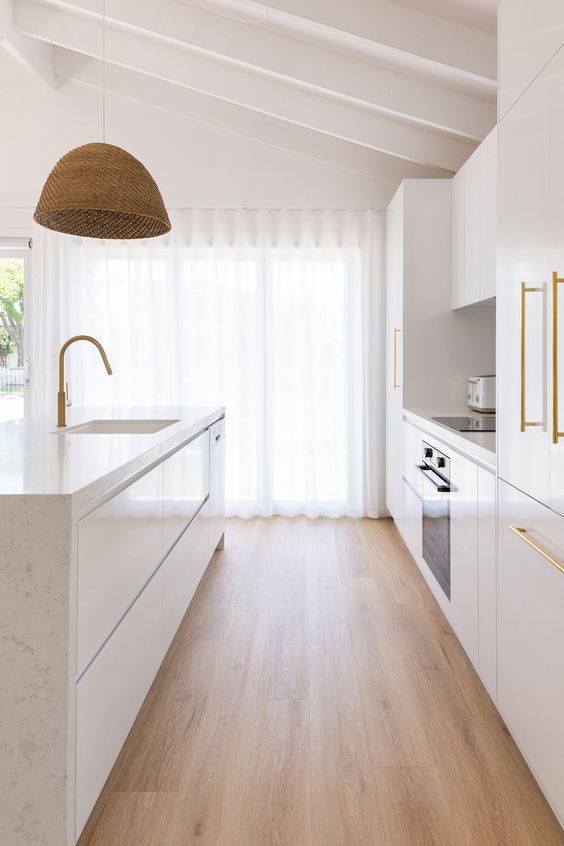 A beautiful modern white kitchen with sleek cabinets, white stone countertops, gold fixtures and a woven pendant lamp