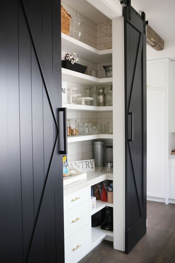 A farmhouse-style pantry with black barn doors, white shelves, and a few storage units is a cool space for any kitchen