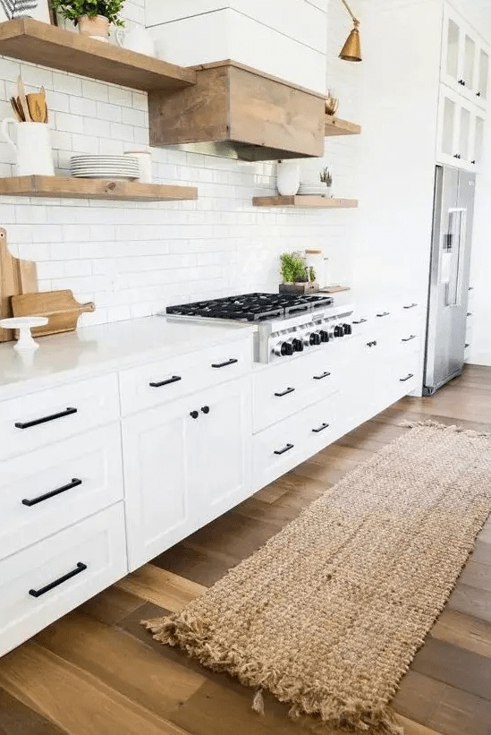 A white modern farmhouse kitchen with shaker-style cabinets, white stone countertops, wooden floating shelves and a wooden hood