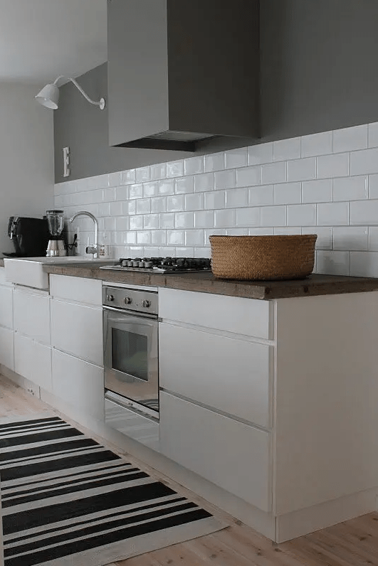 a white Scandinavian kitchen with sleek cabinets, butcher block countertops, a white subway tile backsplash, and a range hood that matches the wall