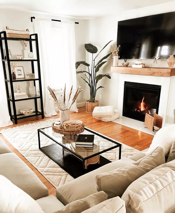a small and inviting living room with a fireplace, neutral seating area, shelf, coffee table, potted plants and pillows