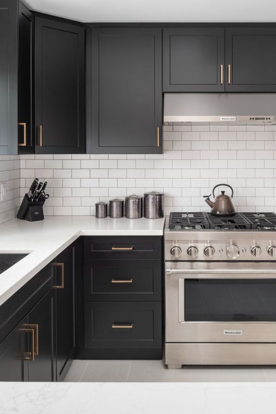 a contrasting kitchen with black cabinets, a white subway tile backsplash and white stone countertops and brass handles