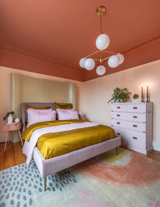 a striking bedroom with a red blanket, a purple bed with mustard linens, a purple dresser, a statement rug and a pink nightstand