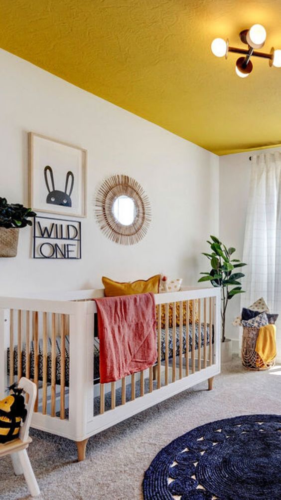 a bright children's room with a mustard-colored blanket, a crib, layered rugs, a stool, a gallery wall and some light pillows