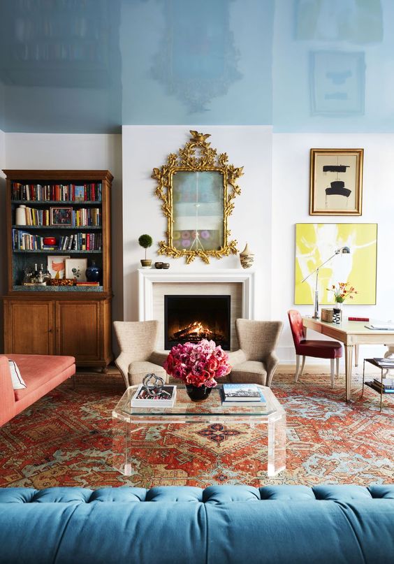 a bright, eclectic living room with a glossy blue ceiling, vintage bookshelf, fireplace, bold printed rug, red daybed and blue sofa