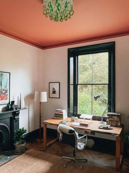 a bright study with a red ceiling, a black fireplace, a vintage desk, a chair, some lamps and artwork, and a green chandelier