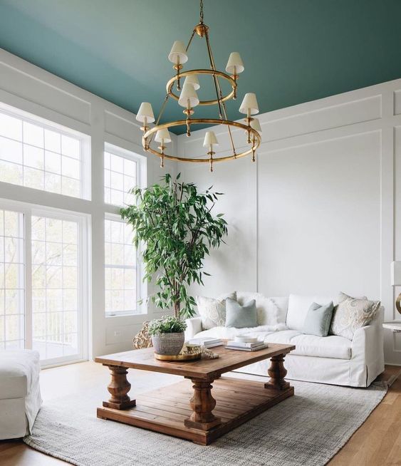 an airy living room with a green blanket, a white sofa, a stained vintage table, a tiered chandelier and a rug