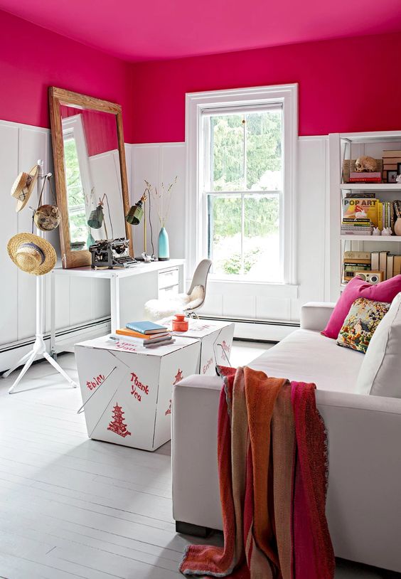 A small, coolly decorated room with a fuchsia ceiling, a desk, a chair, a white sofa and colorful cushions, and a shelf
