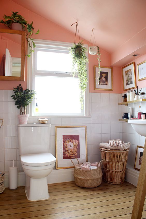 a small and cozy bathroom with a pink sloping ceiling, a sink, a toilet, some baskets and potted plants and decor