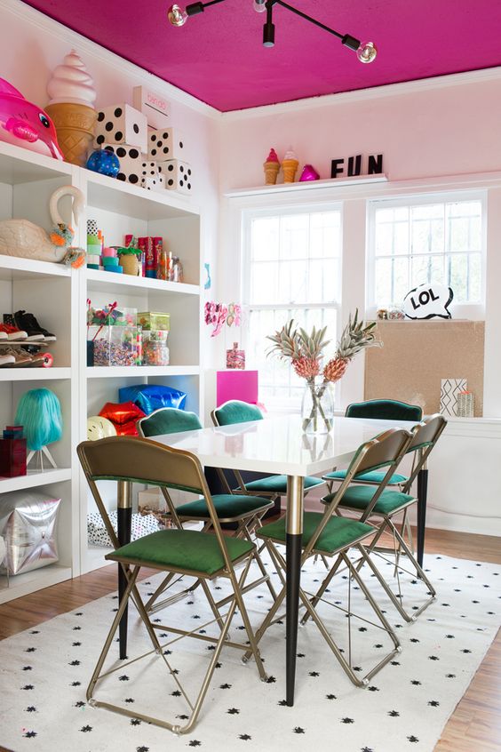 A modern, fun dining room with a fuchsia ceiling, shelf, table, green chairs and lots of whimsical and cool decor