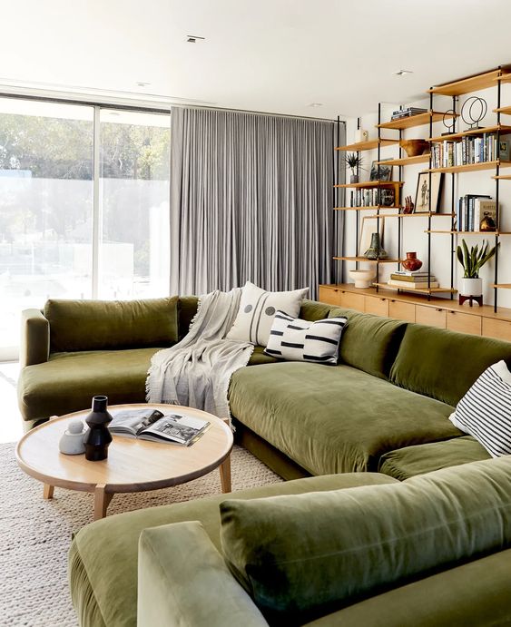 a modern living room with a storage space with open shelves, a large green seating area, a coffee table, gray curtains and printed cushions
