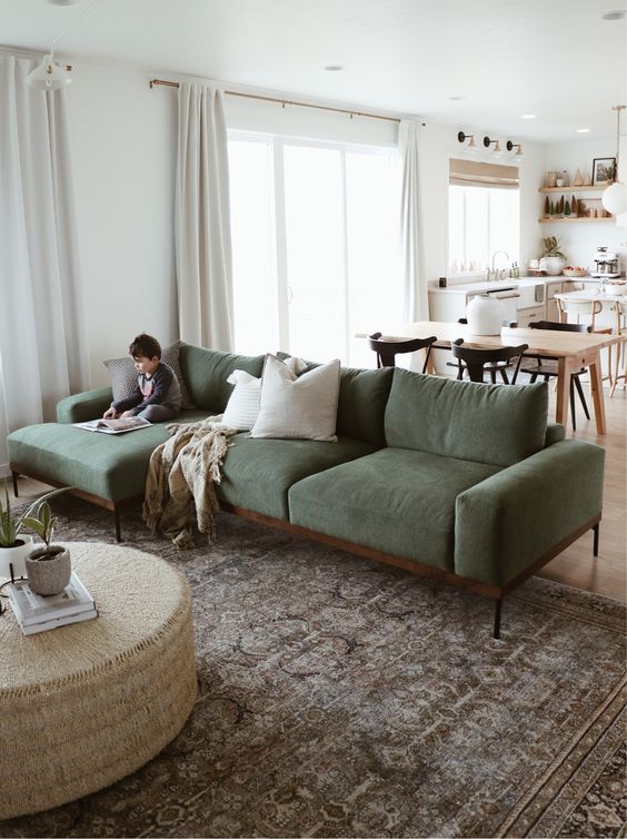 a modern space with a kitchen and dining area and a living room with a green sofa, a coffee table, a rug and neutral curtains