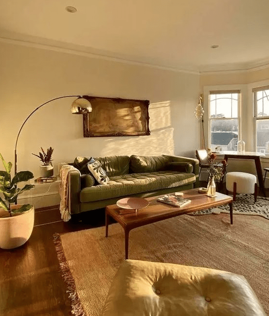 an earthy living room with a green sofa, a yellow stool, a table with some chairs, a low coffee table and some plants