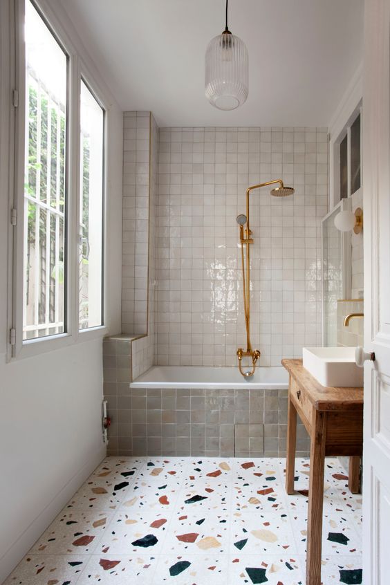 a creative bathroom with a large window, a bath area covered in white and gray zellige tiles, a terrazzo floor, a stained vanity and sink and brass fittings