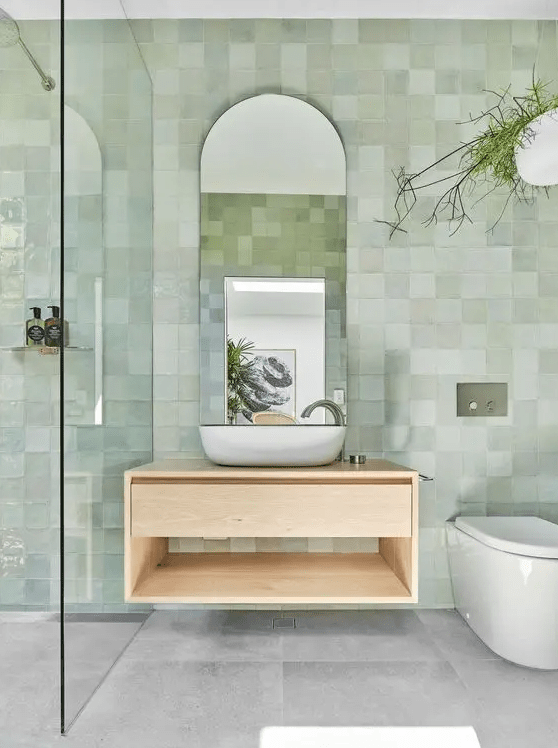 a dreamy and airy bathroom with green zellige tiles on the walls, gray tiles on the floor, a floating vanity and an arched mirror