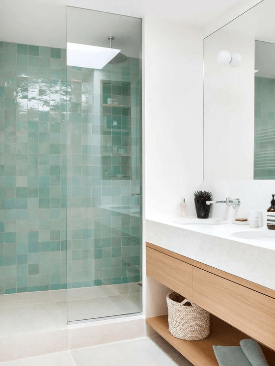 a modern bathroom in neutral tones with a shower area clad in green and turquoise zellige tiles, a light stained vanity and a mirror