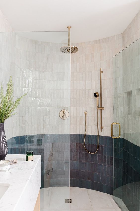 a large, rounded shower area clad in thin navy and white zellige tiles, a marble slab sink, brass hardware and greenery