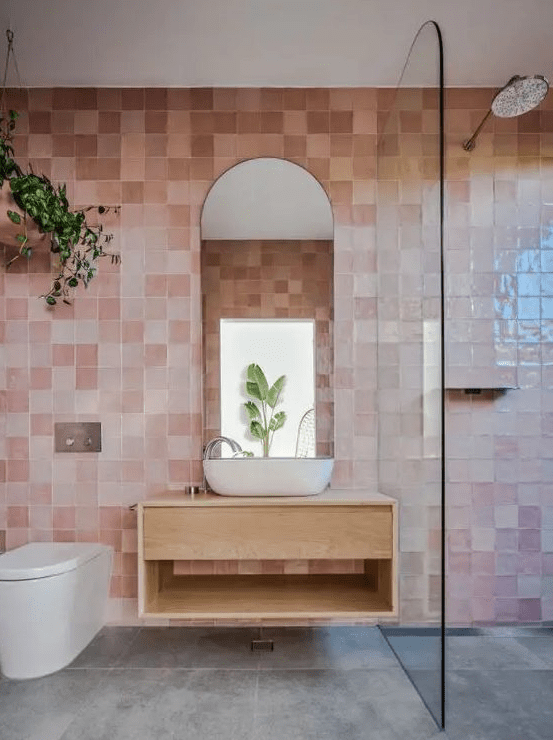 a gorgeous bathroom with pink zellige tiles, a shower, a floating vanity, an arched mirror and some greenery