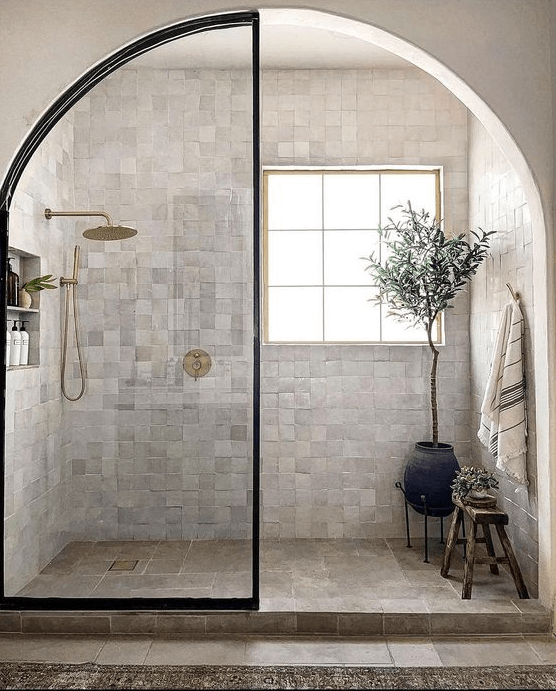 A beautiful shower area with zellige tiles, a window, an arched door and some potted plants