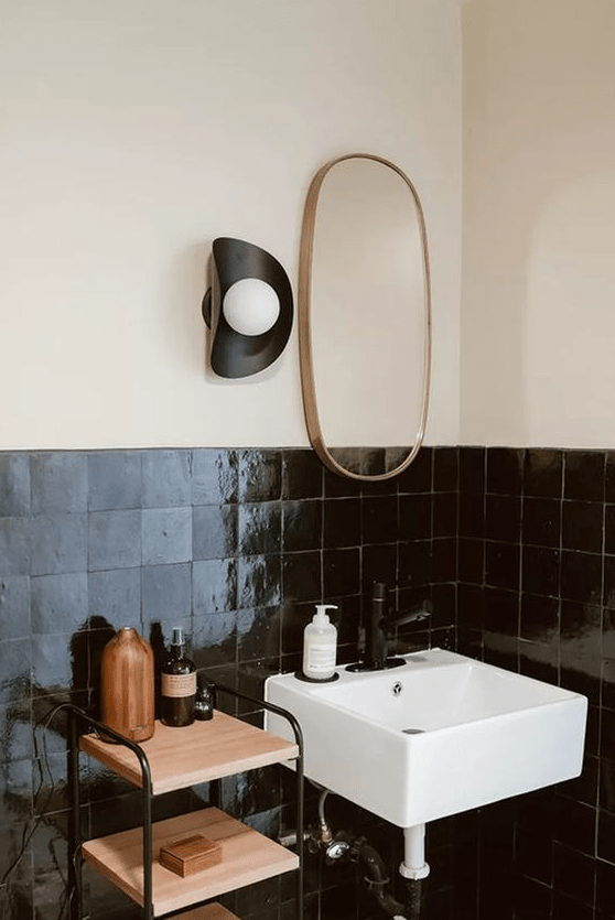 a contrasting bathroom with neutral walls and black zellige tiles, a tiered shelf and a small sink and mirror