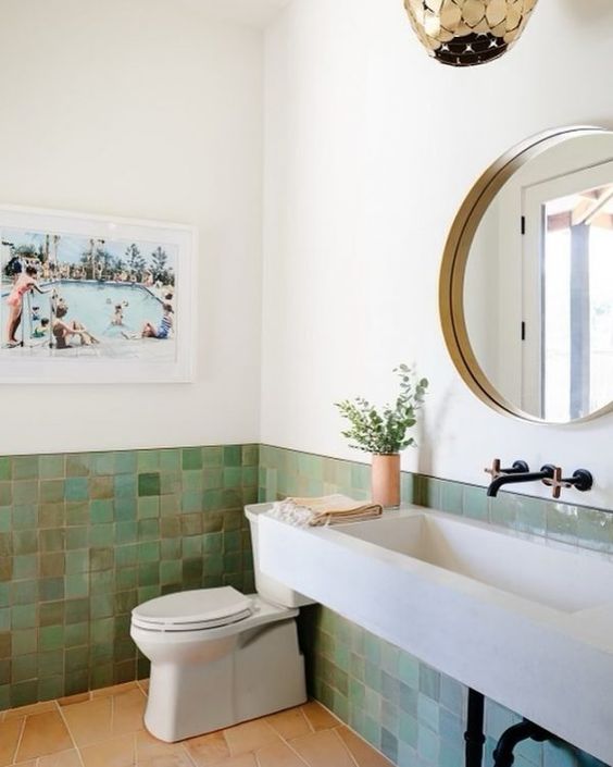a modern bathroom with green zellige tiles, a wall-mounted concrete sink, a toilet, a gold pendant lamp and a round mirror