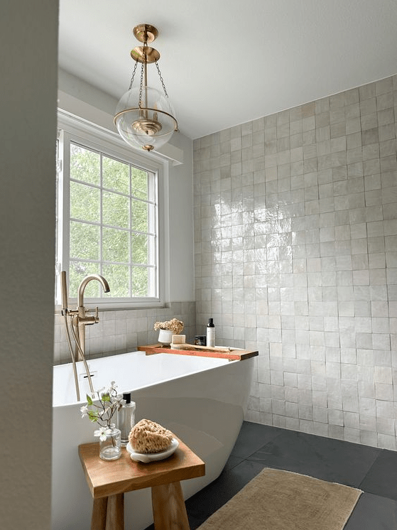 a beautiful modern bathroom with a neutral zellige tile floor, a gray floor, a bathtub, a wooden stool and a pendant lamp
