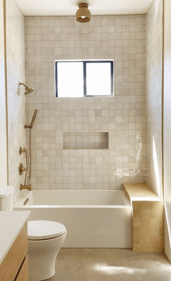 a small neutral bathroom with zellige tiles, a bathtub and a zellige tile bench, brass fixtures and a window