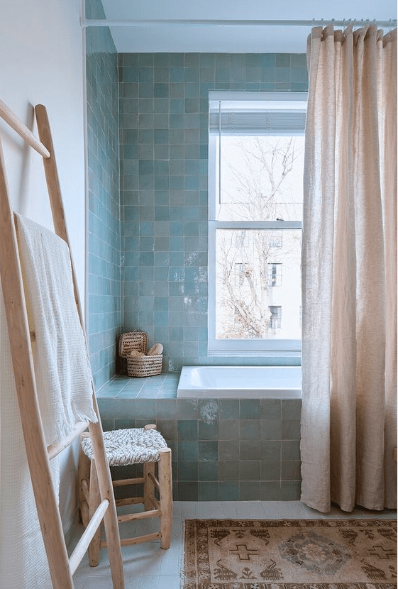 a calm bathroom with green and blue zellige tiles, a bathtub with a pink curtain, a carpet and some wooden furniture