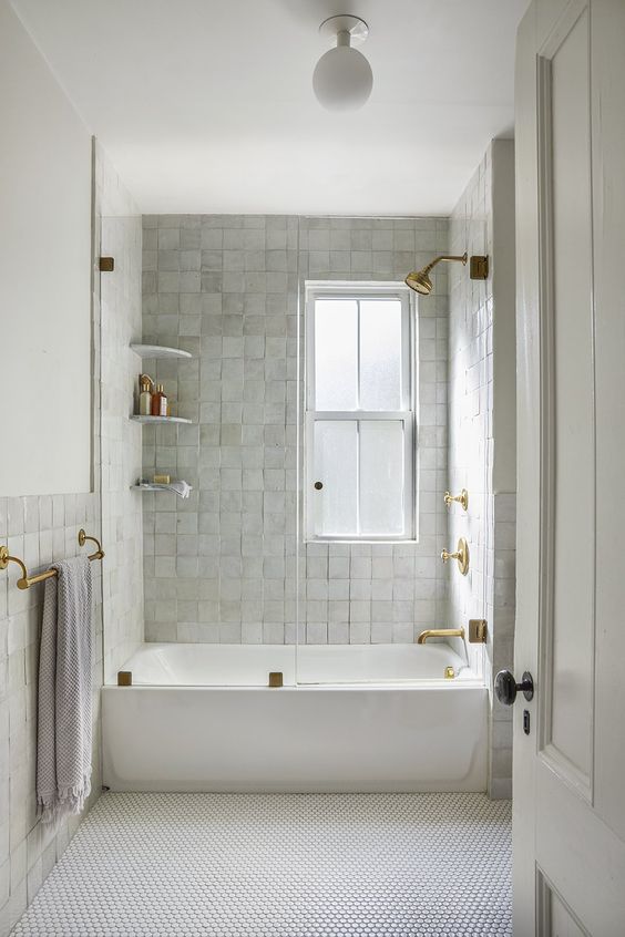 An all-white bathroom made even more striking with zellige and penny tiles, a bathtub, shelves and gold fixtures
