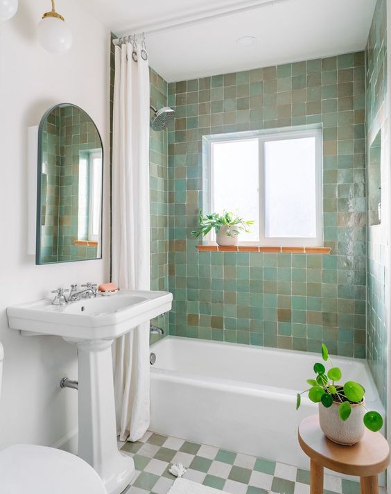 a retro bathroom with green and brown zellige tiles in the tub area, a window, a freestanding bathtub, a stool with a potted plant and a shower curtain