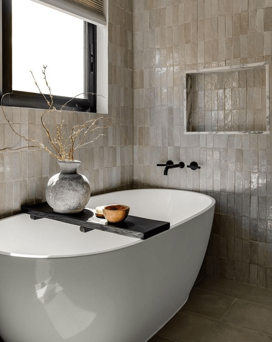 a neutral bathroom with stacked zellige tiles, an oval bathtub, a niche, some branches in a vase and a window