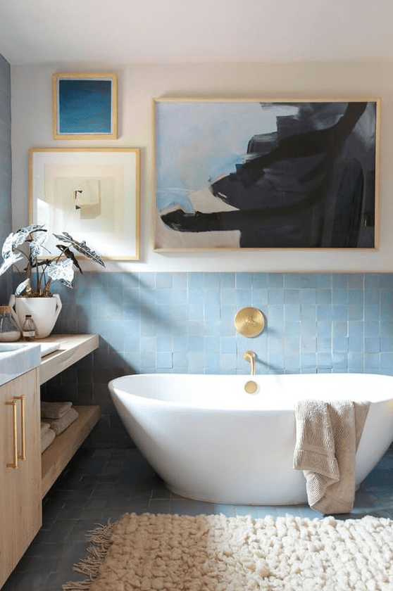 an elegant bathroom with white walls, blue zellige tiles, an oval bathtub, light stained furniture, a gallery wall and gold fixtures