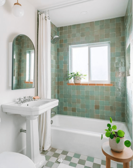 a small and pretty bathroom with green and beige zellige tiles, a checkered floor, a freestanding bathtub and some potted plants
