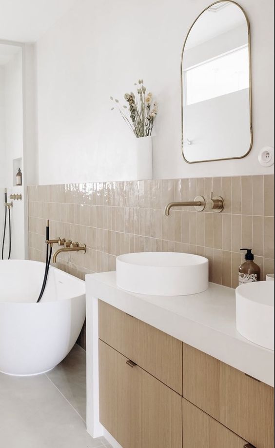 a modern, rustic bathroom with white walls, light brown zellige tiles, a built-in vanity with drawers, a bathtub, round sinks and a mirror