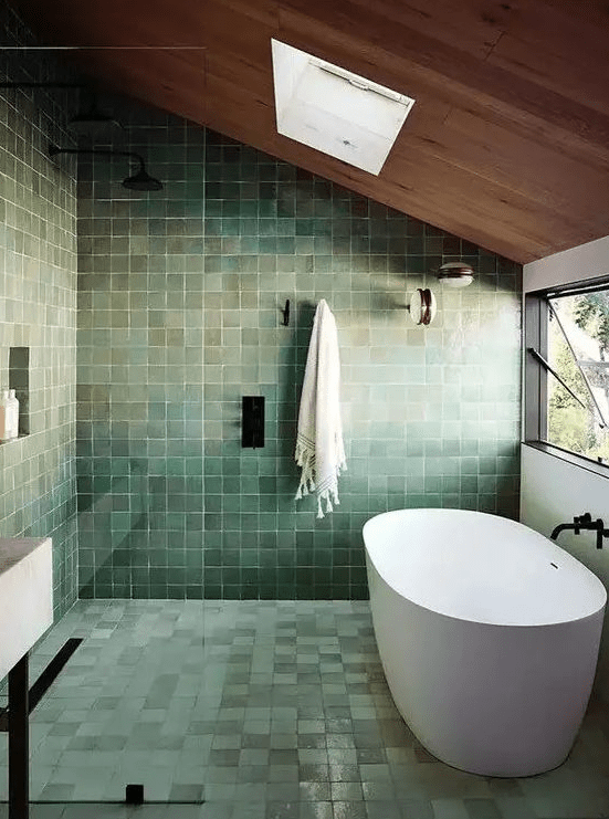 A striking attic bathroom, complete with green zellige tiles, with a skylight and window and black fixtures for a modern feel