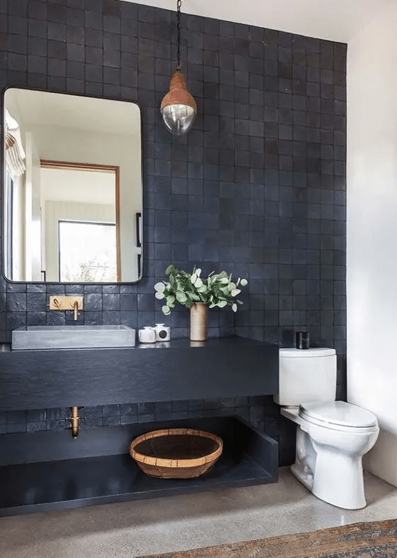 a stylish bathroom with a wall made of black zellige tiles, a black vanity unit, a rectangular mirror, a hanging lamp and a gray stone sink