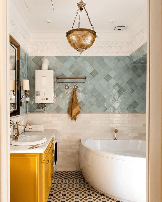 a striking bathroom with gray and green zellige tiles, a corner bathtub, a yellow vanity and a brass pendant lamp