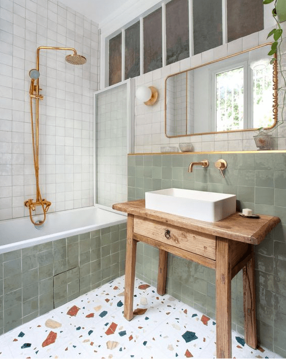 a striking bathroom with white and green zellige tiles, a terrazzo floor, a stained vanity and gold fixtures