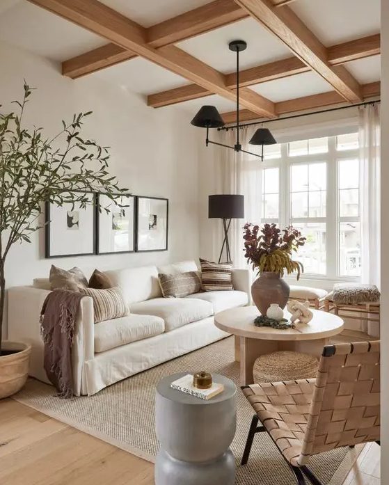 a light-filled, earthy living room with exposed beams, a white sofa, stools and a wicker chair, a gray side table and a round coffee table