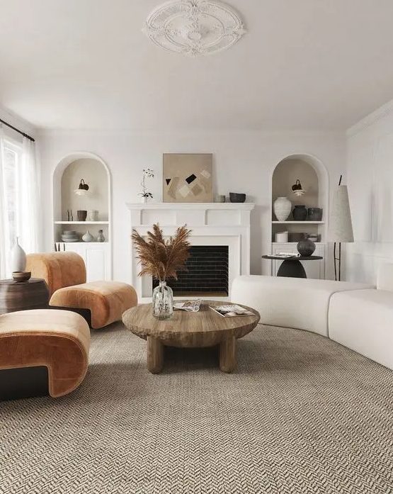 a modern, earthy living room with a fireplace, rust-colored chairs, a neutral curved sofa, a low coffee table and niches with decor