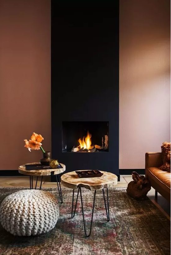 a sophisticated, modern living room with a rust-colored wall and a black fireplace, a leather sofa, wooden slice tables and a knitted ottoman