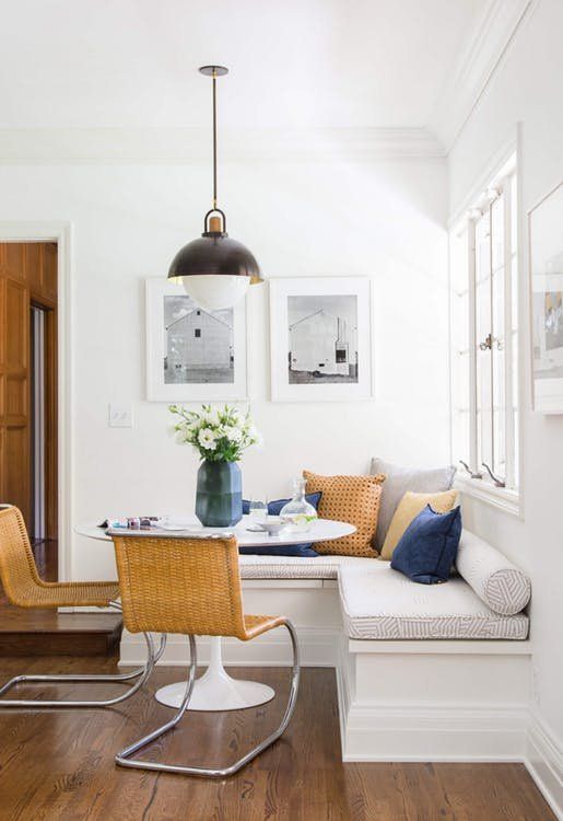 a chic, modern breakfast nook with a corner bench and cushions, a round table, woven chairs, a vintage pendant lamp and some artwork