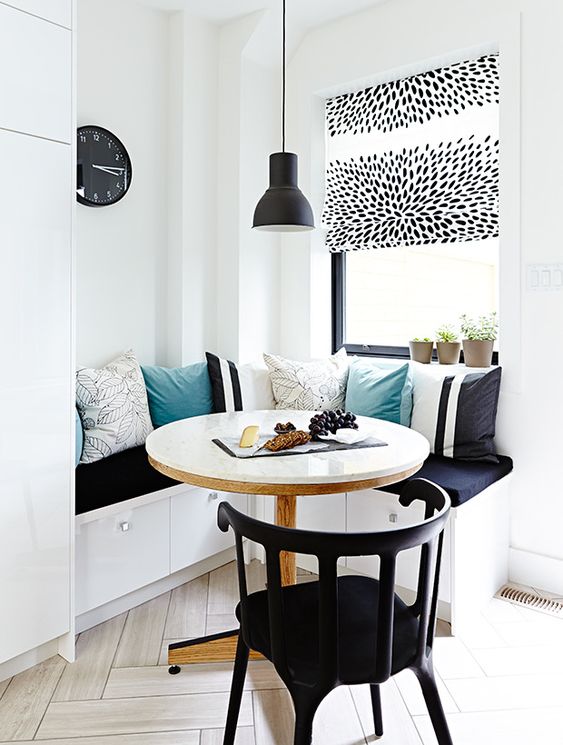 a contrasting breakfast nook with a corner bench and lots of cushions, a round table and a black chair, a black pendant lamp and a clock