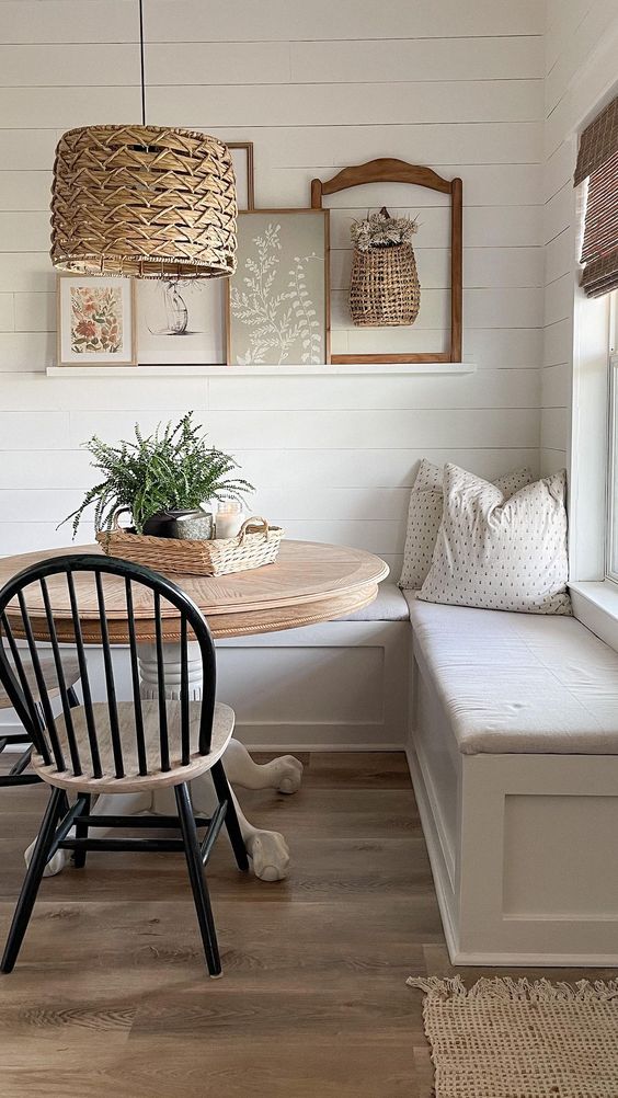 a cottage breakfast nook with corner seating and cushions, a stained round table, a wicker pendant lamp and a gallery wall