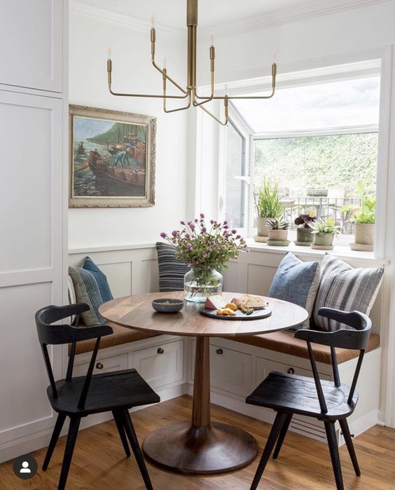 a farmhouse-style breakfast nook with a corner bench, a stained table and black chairs, a chandelier and potted plants, and a piece of art