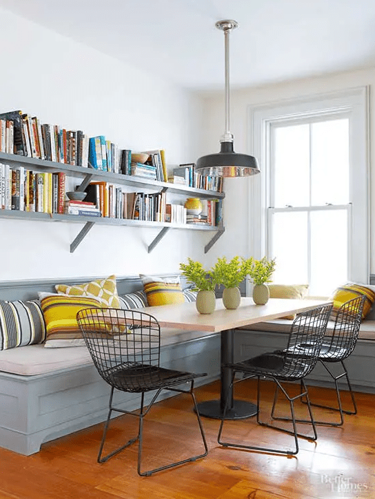 A farm dining area with a gray bench seat with colorful cushions, a table on a metal leg, open shelves and black metal chairs